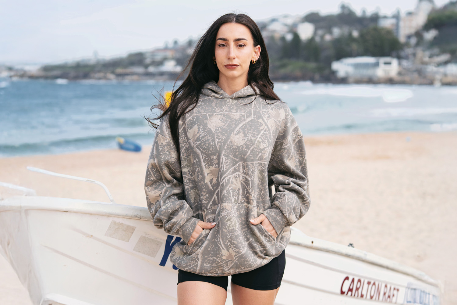 girl wearing a Deadwood Tree Camo hoodie standing next to a boat on the beach